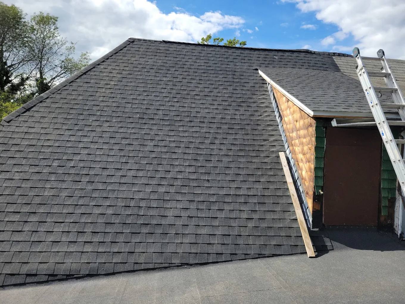 Newly installed black architectural shingles on a hip roof, with a ladder at the dormer